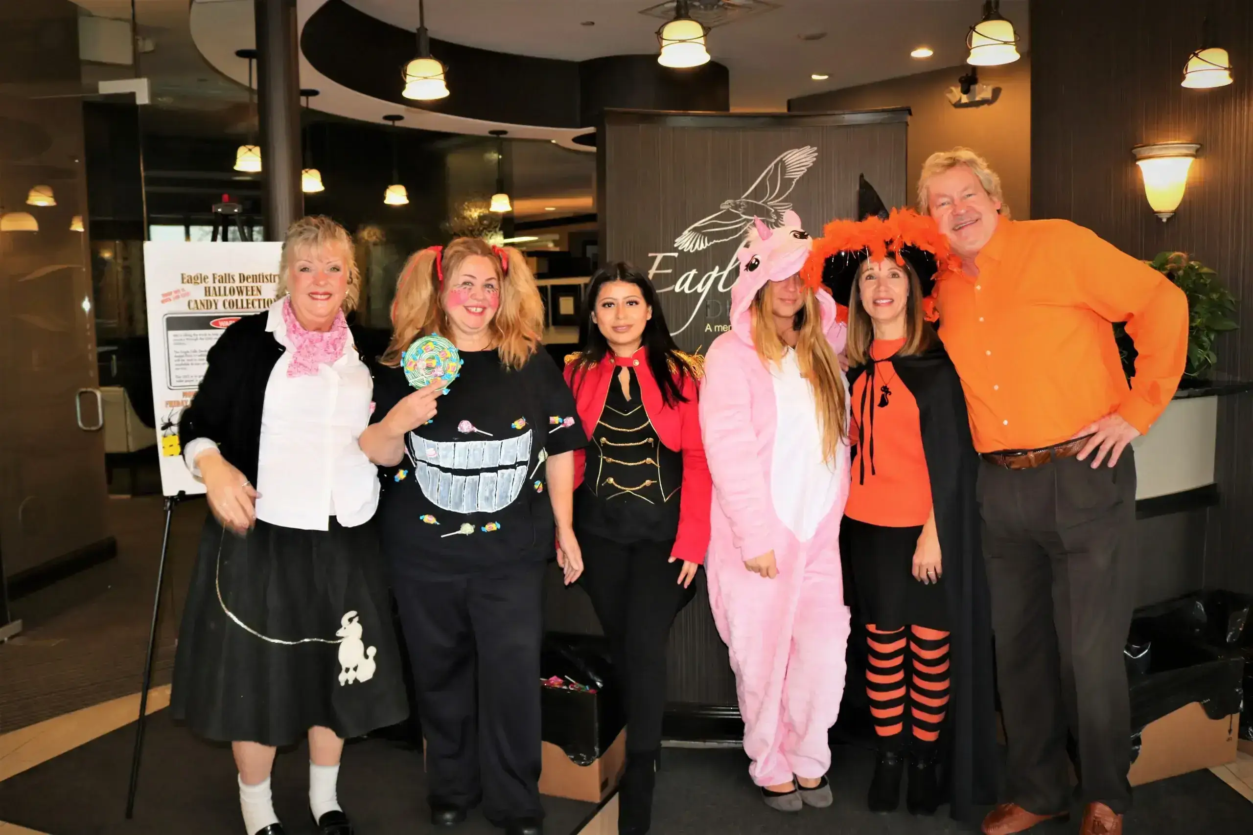 Group of six people in a warmly lit room, dressed in playful Halloween costumes including a poodle skirt, unicorn onesie, and vampire cape, smiling and festive.