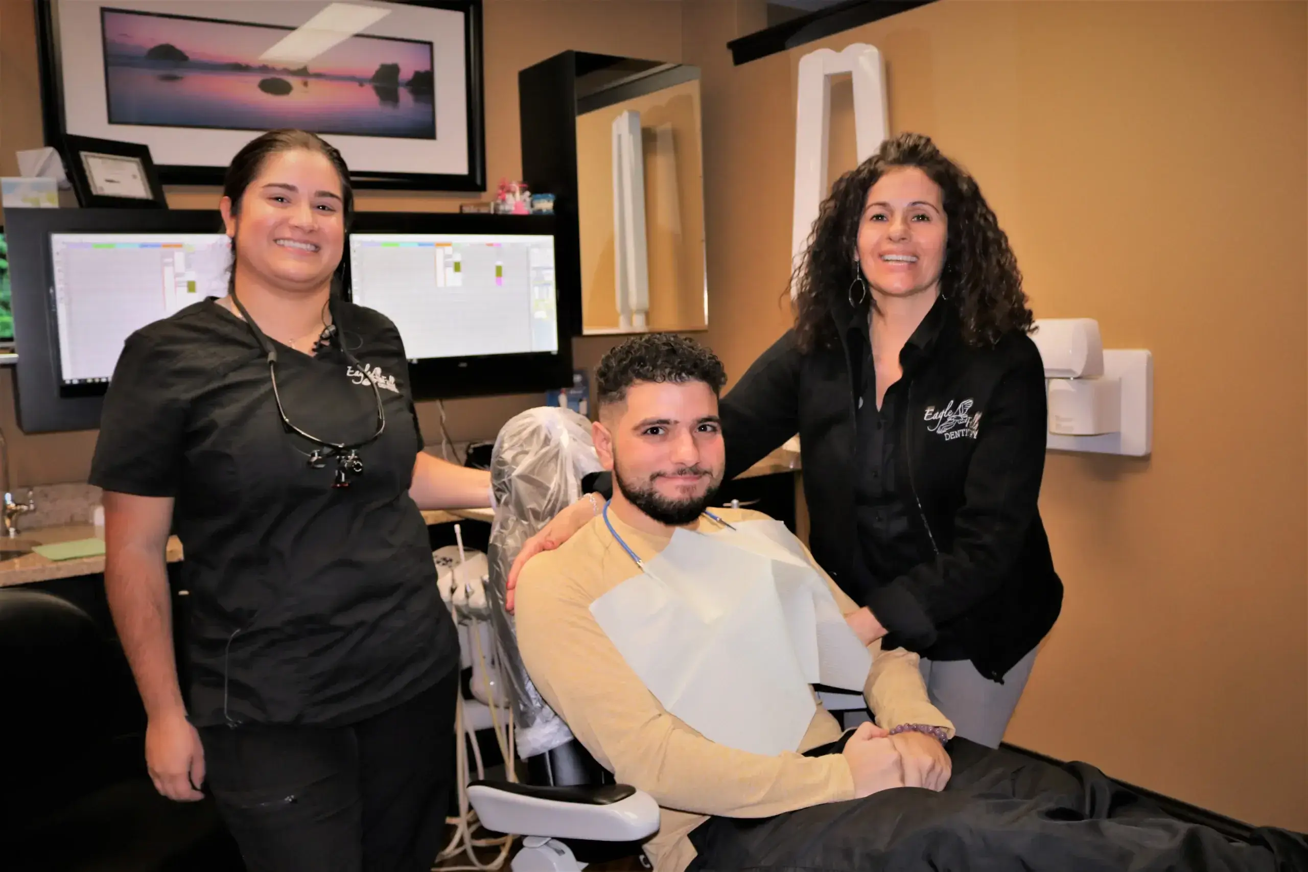 A dentist office setting with a smiling patient in a dental chair, flanked by two dental staff members. Monitors display dental software behind them.