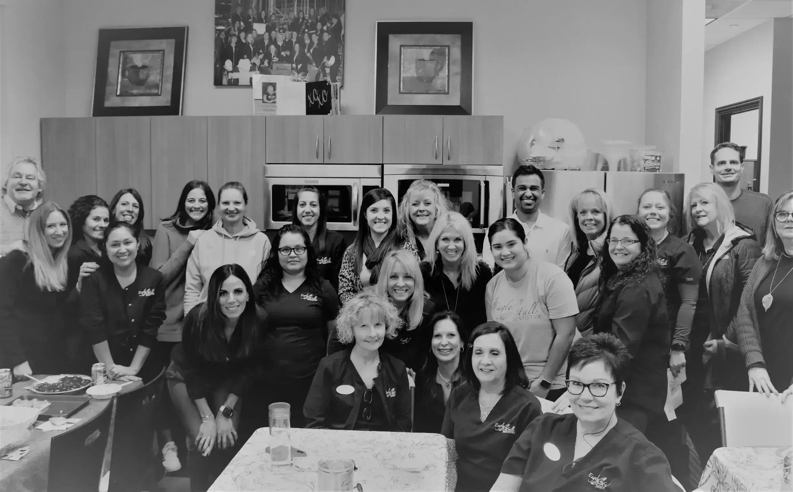 A group of people smiling in an office break room with cabinets and art on the walls. The mood is friendly and relaxed. Some individuals wear matching uniforms.