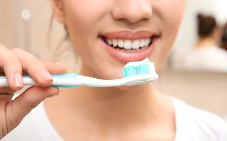 A person smiling with white teeth, holding a toothbrush with blue bristles and toothpaste. The mood is fresh and clean, conveying dental hygiene.