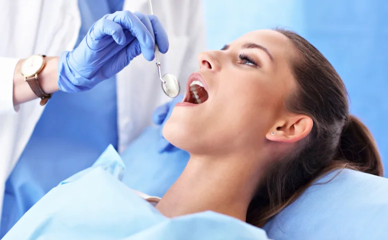 A dentist wearing blue gloves examines a female patient's open mouth using a dental mirror. The patient looks calm, surrounded by a bright, clinical setting.